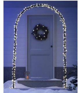 Arche de Noël lumineuse installée en extérieur devant une porte d’entrée, au sol enneigé, avec un sapin de Noël à gauche